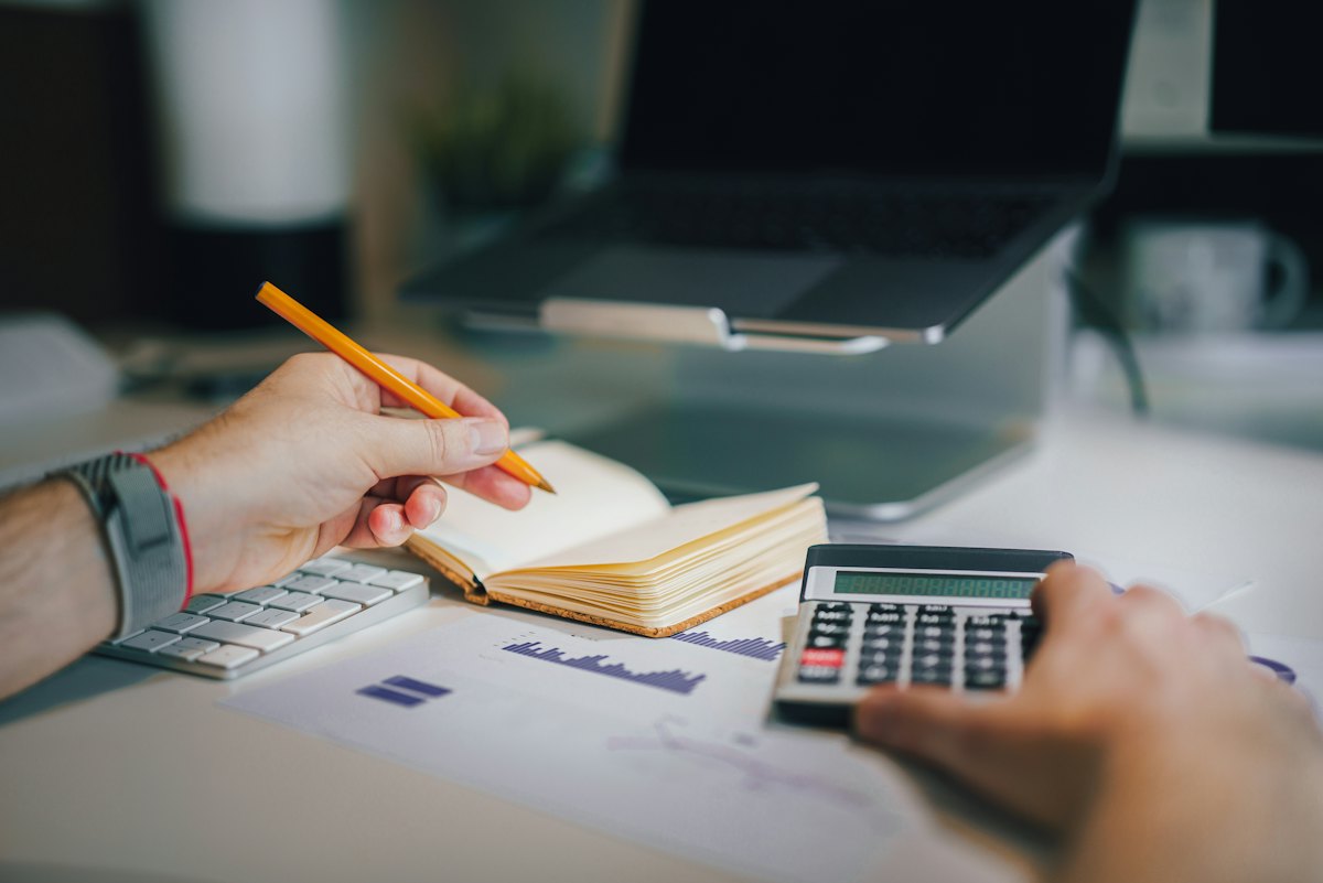 Mains sur une calculatrice et un carnet ouvert, devant un ordinateur portable